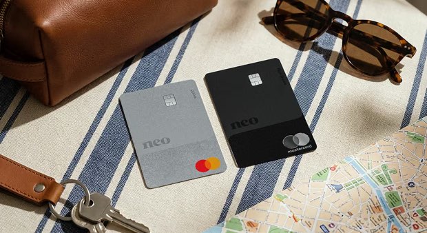 Two Neo Mastercard credit cards, silver and black, on a striped cloth beside sunglasses, keys and a map